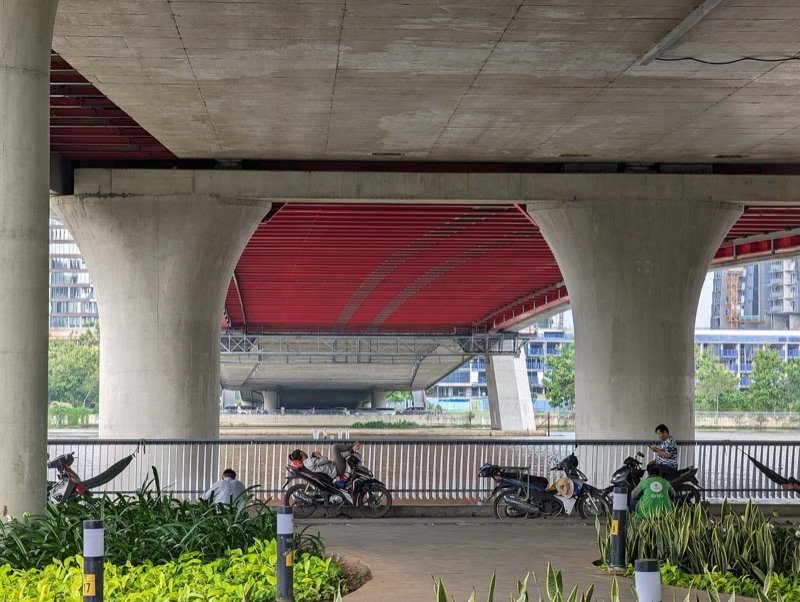 Người dân nghỉ ngơi trên võng và xe máy dưới gầm cầu Ba Son, bên lối đi bộ ven sông có cây xanh