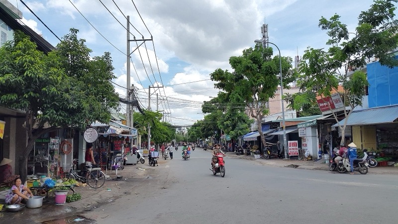 Con phố nhỏ ở Quận 2 với hàng quán hai bên, người bán rau ngồi bên lề đường, xe máy qua lại dưới hàng cây